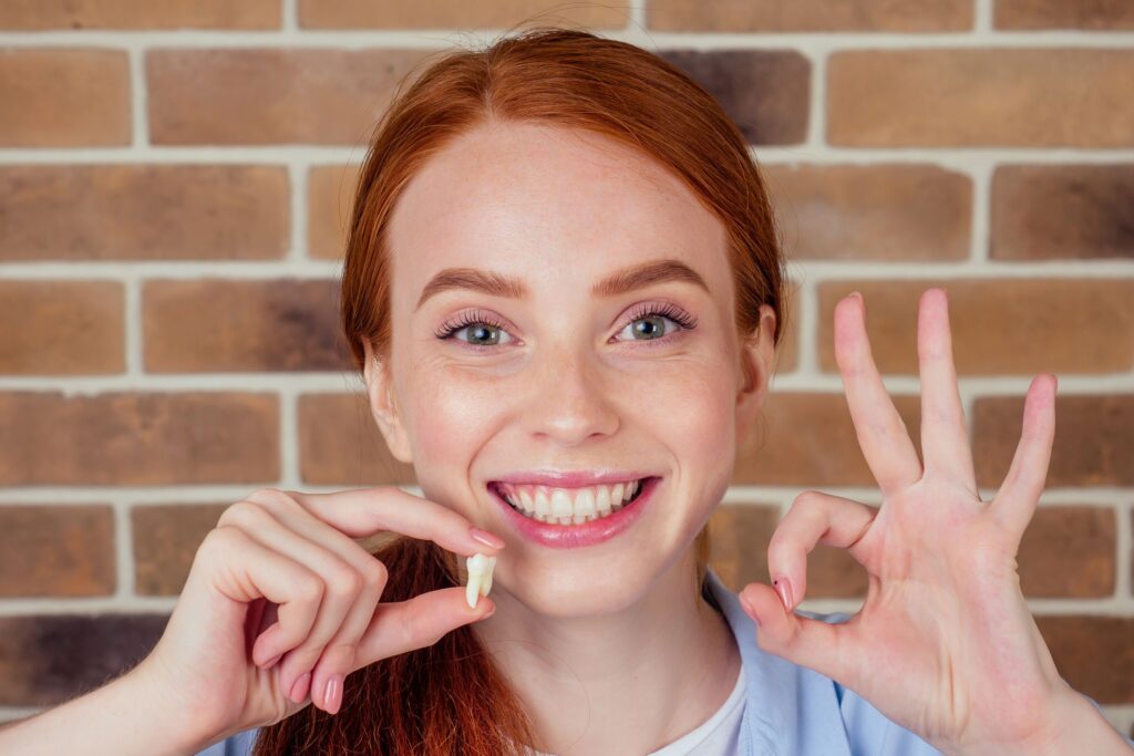 Woman with red hair holding extracted tooth and making 'ok' sign with other hand